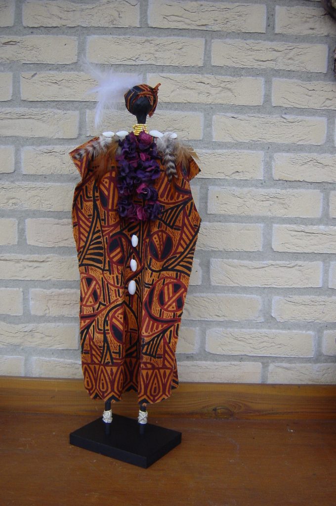 AFRIKAANSE VROUW van stof Het lichaam is bekleed met een Afrikaans stofje en versierd met schelpen, veertjes, geverfde tuinkrullen, kraaltjes en wat touw voor de enkels. De hoogte van de pop is ongeveer 56 cm
