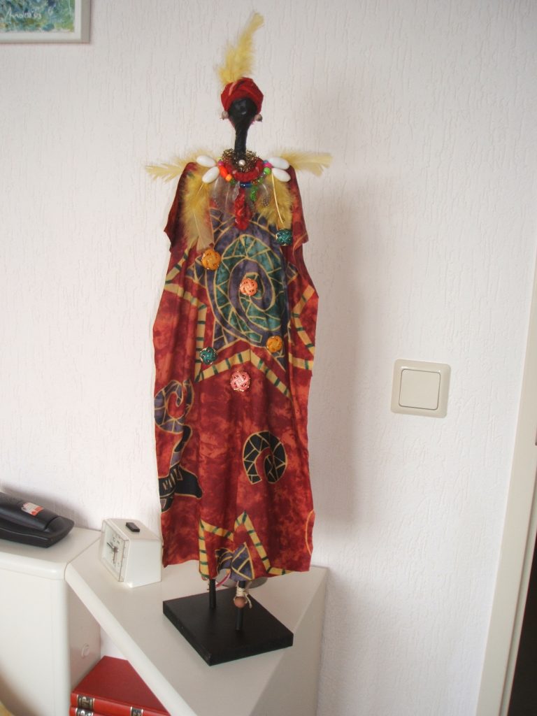 AFRIKAANSE VROUW Het lichaam is met een Afrikaans stofje bekleed en bewerkt met textielverharder. Versierd met kralen, veren en knopen. De hoogte van de pop is ongeveer 60 cm