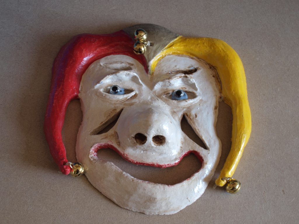 Masker Harlekijn, geboetseerd, witte klei nr 478 met grove chamotte. Gestookt 960° C. Bewerkt met kleurpigmenten geel/groen/rood. Transparant mat geglazuurd 1060° C. Afgewerkt met belletjes en glazen ogen.